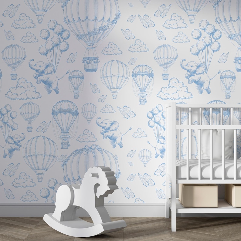 May include: A white nursery wall with a blue and white wallpaper featuring hot air balloons, clouds, butterflies, and elephants. A white rocking horse and a white crib are in the foreground.