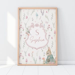 Op de afbeelding: Een roze en witte bloemenprint met een konijn dat een gieter vasthoudt. De print heeft een roze wapenschild met de letter "S" en de naam "Sophia" in een handschrift.