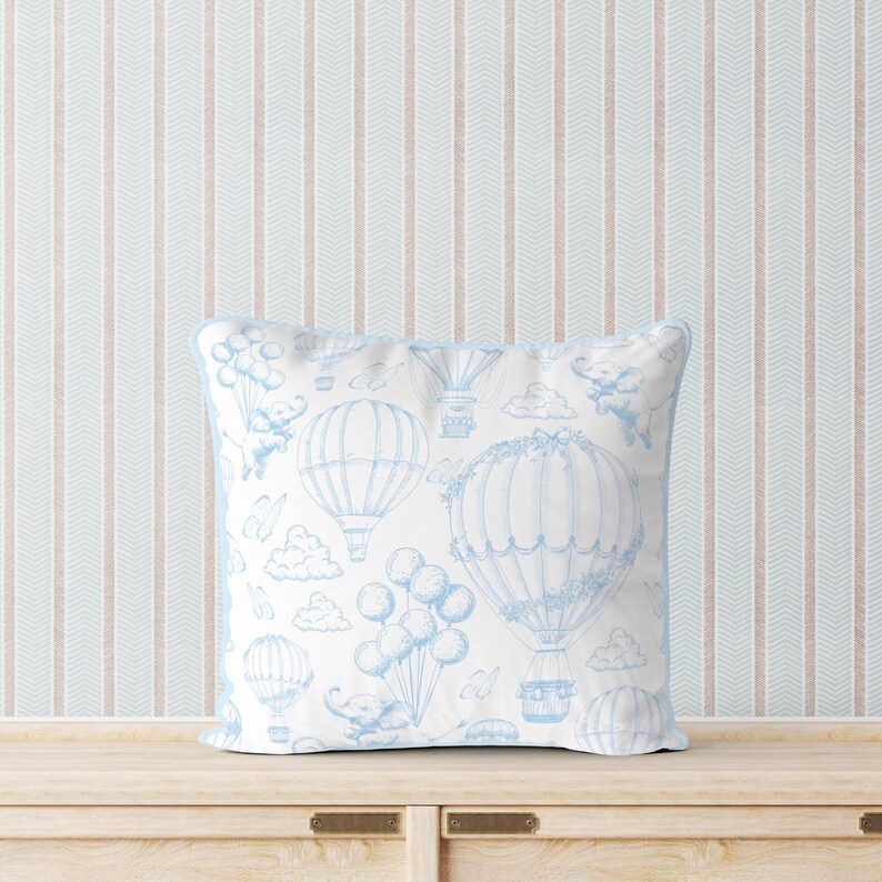 May include: A white throw pillow with a blue and white pattern of hot air balloons, clouds, and elephants. The pillow is on a light wood surface with a light blue and pink striped wallpaper behind it.