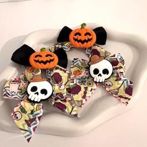 May include: Three hair bows featuring Halloween-themed patterns with ghosts, skulls, pumpkins, and brooms. Two bows are topped with white skull charms wearing brown hats, and one bow features an orange jack-o'-lantern charm with black accents.