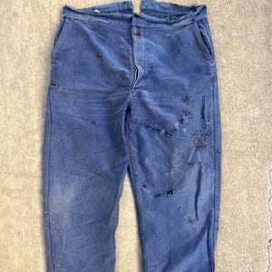 Puede incluir: Pantalones de trabajo azules descoloridos con desgaste visible. Los pantalones tienen bragueta con botones, múltiples bolsillos y áreas desgastadas. La tela muestra signos de edad y uso, con decoloración y parches. Estilo vintage.