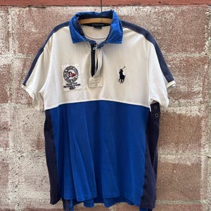 May include: A short-sleeved polo shirt in white and blue, featuring a navy blue collar and accents. The shirt has a patch with text and a logo on the chest, and a small embroidered logo of a person on a horse. The shirt is hanging on a wooden hanger.