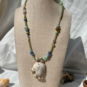 May include: A handmade necklace featuring a large, textured seashell pendant. The necklace has a green cord with various colorful beads in shades of blue, green, and gold. The seashell is a natural, off-white color. The necklace is displayed on a neutral-toned mannequin.