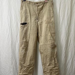 May include: Tan work pants with visible wear and tear, including a large hole on the left leg and patches. The pants have a button closure, belt loops, and a small red and white label near the right pocket. The fabric appears worn and faded.