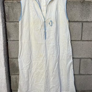 May include: A sleeveless white dress with blue trim and embroidered details. The dress features a collared neckline, a button-down front, and a blue embroidered emblem. The dress is hanging on a wooden hanger against a gray brick wall.