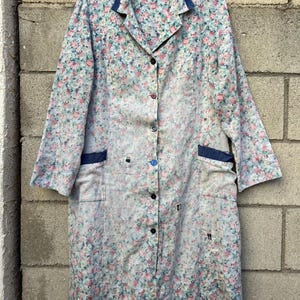 May include: A long-sleeved floral print coat with a button-up front, dark blue trim, and two front pockets. The coat features a pattern of small pink, blue, and green flowers on a light background.
