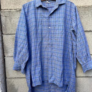May include: A blue plaid shirt with long sleeves and a collar, suspended from a wooden hanger. The shirt features a button-down front and a high-low hem. The plaid pattern incorporates blue, grey, and red hues.