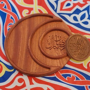 May include: Wooden decorative set featuring three pieces of varying sizes. The largest piece is a crescent shape, with two smaller circular pieces. The pieces have Arabic calligraphy carved into them. The background is a colorful patterned fabric.