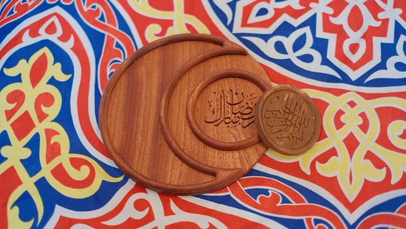 May include: Wooden decorative set featuring three pieces of varying sizes. The largest piece is a crescent shape, with two smaller circular pieces. The pieces have Arabic calligraphy carved into them. The background is a colorful patterned fabric.