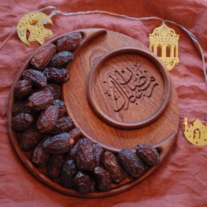 May include: A wooden crescent-shaped serving tray filled with dates, with a circular inset featuring Arabic calligraphy. Gold-colored moon and lantern string lights are arranged around the tray, set against a red-brown fabric.