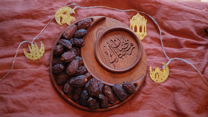 May include: A wooden crescent-shaped serving tray filled with dates, with a circular inset featuring Arabic calligraphy. Gold-colored moon and lantern string lights are arranged around the tray, set against a red-brown fabric.