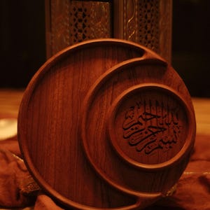 May include: A wooden serving tray with a circular design and carved Arabic calligraphy. The tray is a warm brown color and has a smooth finish. A gold-colored lantern is in the background.