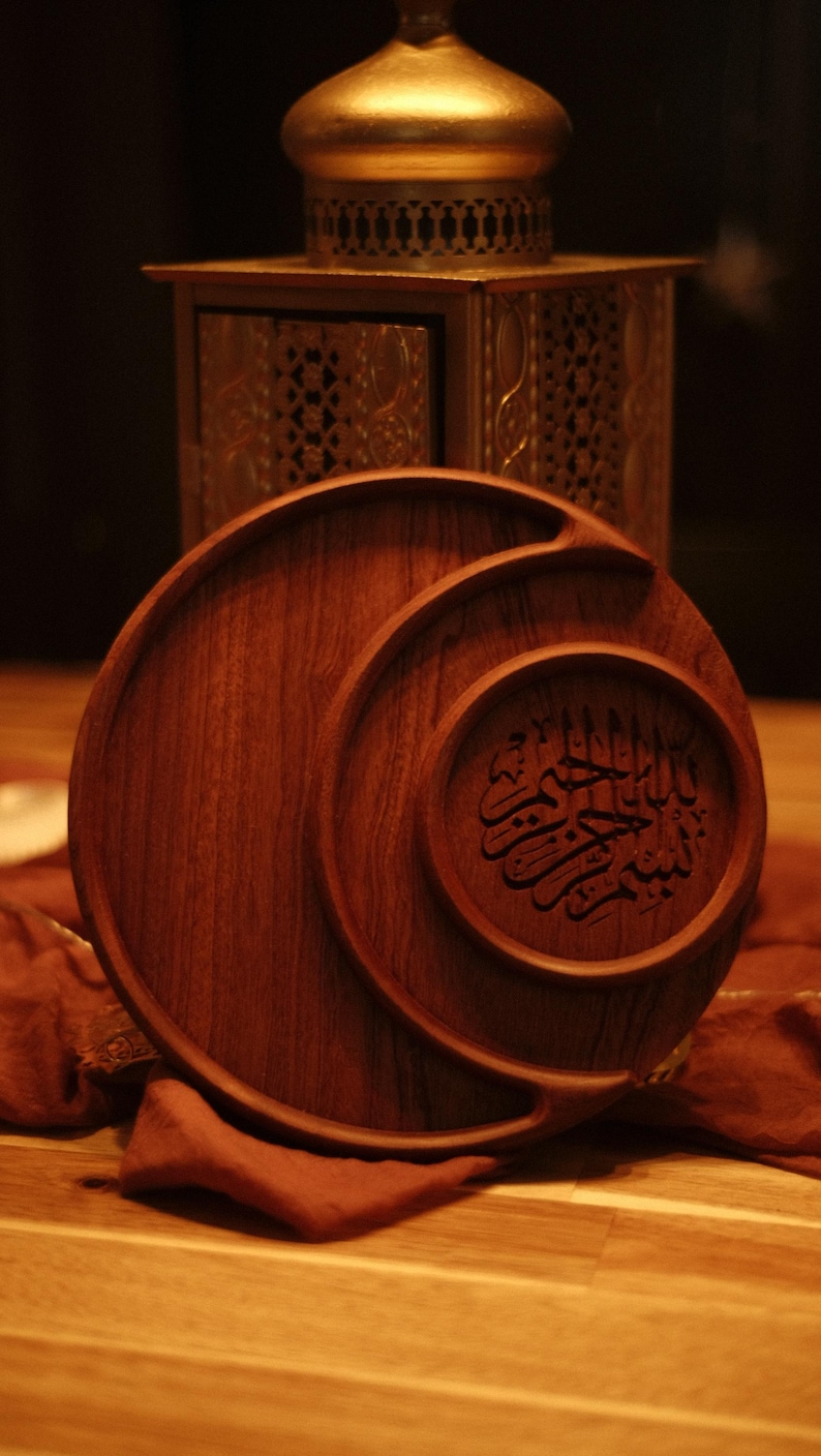 May include: A wooden serving tray with a circular design and carved Arabic calligraphy. The tray is a warm brown color and has a smooth finish. A gold-colored lantern is in the background.