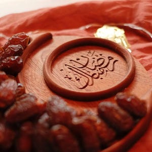 May include: A wooden serving tray with a crescent moon shape, filled with dates. A smaller round wooden piece with Arabic calligraphy is in the center. Decorative lights in the shape of moons and stars are in the background.