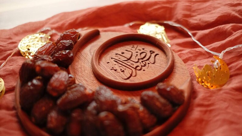 May include: A wooden serving tray with a crescent moon shape, filled with dates. A smaller round wooden piece with Arabic calligraphy is in the center. Decorative lights in the shape of moons and stars are in the background.