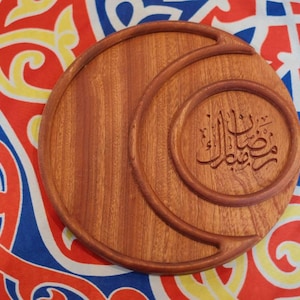 May include: A round, wooden serving tray with a crescent moon design and Arabic calligraphy. The tray is a warm brown color and sits on a colorful patterned fabric with red, yellow, and blue hues. The calligraphy is in a circular frame.