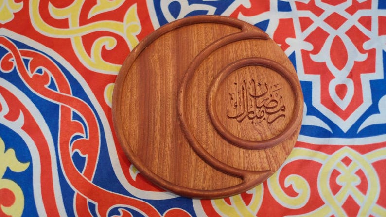 May include: A round, wooden serving tray with a crescent moon design and Arabic calligraphy. The tray is a warm brown color and sits on a colorful patterned fabric with red, yellow, and blue hues. The calligraphy is in a circular frame.
