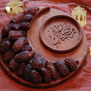 May include: A wooden crescent-shaped serving tray filled with dates, alongside a round wooden dish with Arabic calligraphy. Gold-colored decorative lights in the shape of moons and lanterns are strung around the tray, all set against a red background.