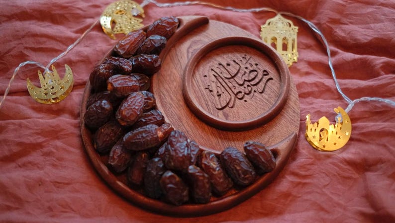 May include: A wooden crescent-shaped serving tray filled with dates, alongside a round wooden dish with Arabic calligraphy. Gold-colored decorative lights in the shape of moons and lanterns are strung around the tray, all set against a red background.