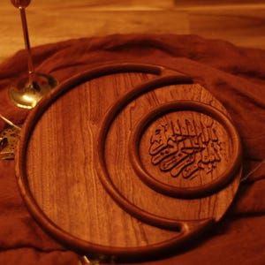 May include: A wooden serving tray with a crescent moon design and an inner circular tray with Arabic calligraphy. The tray is made of a warm-toned wood and sits on a reddish-brown fabric. Decorative candle holders and string lights are in the background.