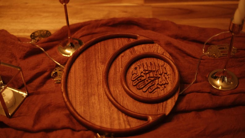 May include: A wooden serving tray with a crescent moon design and an inner circular tray with Arabic calligraphy. The tray is made of a warm-toned wood and sits on a reddish-brown fabric. Decorative candle holders and string lights are in the background.