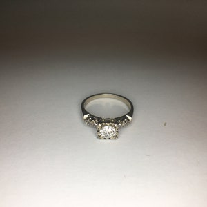 以下が含まれることがあります： 中央に大きく丸い透明なダイヤモンド、バンドに小さなダイヤモンドがついた銀の婚約指輪です。