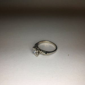以下が含まれることがあります： 中央に丸い透明なダイヤモンドが1つ付いた銀製の婚約指輪。バンドは中央の石の両側に小さなダイヤモンドがセットされています。