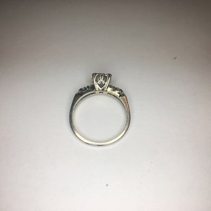 以下が含まれることがあります： 中央に丸いダイヤモンド、バンドに小さなダイヤモンドがついたシルバーの婚約指輪です。