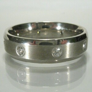 May include: A silver-colored ring with a brushed metal finish. The ring features several small, round, clear gemstones evenly spaced around the band. The ring is displayed on a reflective surface.