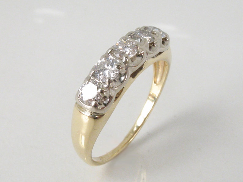 Puede incluir: Un anillo de banda de oro con una fila de cinco diamantes transparentes engastados en un engaste de metal blanco.
