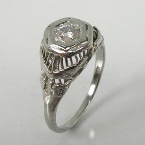 May include: A silver ring with a delicate filigree design and a single round clear stone in the center.