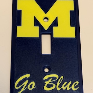 May include: A navy blue light switch cover with a yellow "M" logo and the words "Go Blue". The cover has a rectangular shape with a single switch opening and two screw holes.