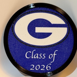 Può includere: Un emblema rotondo blu e bianco con la scritta "Class of 2026". Il design presenta una "G" bianca all'interno di un cerchio blu, circondato da un bordo nero. L'area blu ha un effetto glitterato.
