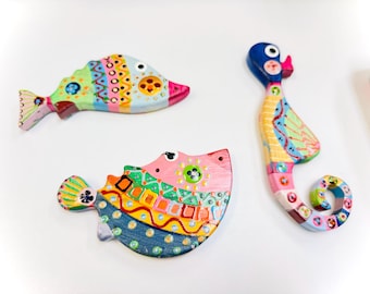 May include: Three colorful wooden fish ornaments, painted in a patchwork style with bright colors and patterns. The fish are a seahorse, a pufferfish, and a fish with a long tail fin.