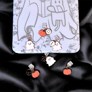 May include: A selection of Halloween-themed phone charms. The charms include a white ghost, an orange pumpkin, and a gold-coloured initial. The charms are attached to a clear plastic clip. The background is a light blue phone case with a floral design.
