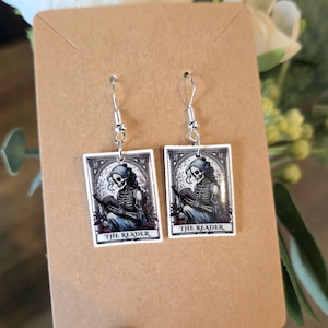 May include: A pair of silver-toned dangle earrings. Each earring features a rectangular pendant with a black and white illustration of a skeleton reading a book. The text "THE READER" is printed at the bottom of each pendant. The earrings are displayed on a brown card.