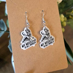 May include: A pair of dangle earrings with a silver hook. Each earring features a white, rectangular charm with a black and white illustration of a woman reading a book. The text "MY WEEKEND IS ALL BOOKED" is printed on each charm.
