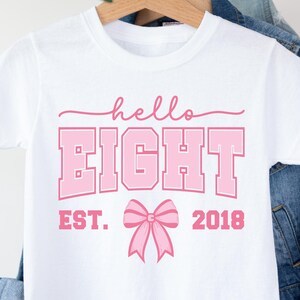 Op de afbeelding: Wit T-shirt met de tekst "hello EIGHT" in roze, een roze strik en de tekst "EST. 2018". Het T-shirt wordt gepresenteerd op een houten kleerhanger.