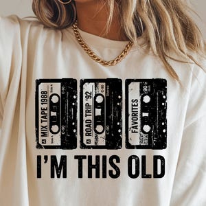 May include: Ivory-colored t-shirt featuring three cassette tape designs with the text "MIX TAPE 1988", "ROAD TRIP '92", and "FAVORITES". Below the tapes, the text reads "I'M THIS OLD".
