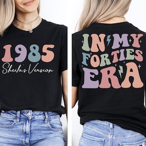 Op de afbeelding: Zwart T-shirt met een retro-ontwerp. De voorkant van het shirt zegt "1985 Sheila's Version". De achterkant van het shirt zegt "In My Forties Era" met bliksemschichten.
