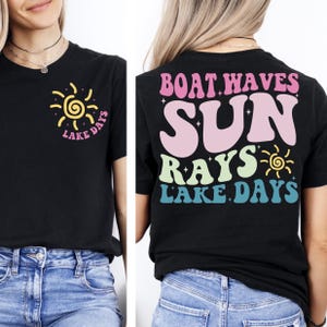 Op de afbeelding: Zwart T-shirt met een retro design met een gele en roze zonnegrafiek en de woorden "Lake Days". De achterkant van het shirt toont de woorden "Boat Waves Sun Rays Lake Days" in een kleurrijk, golvend lettertype.