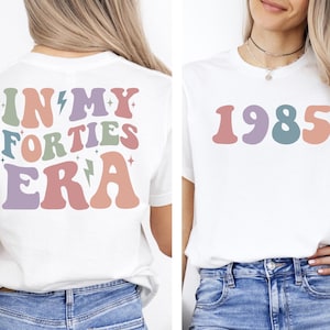 Op de afbeelding: Wit T-shirt met een kleurrijke afbeelding op de achterkant die "In My Forties Era" zegt en een kleurrijke afbeelding op de voorkant die "1985" zegt.