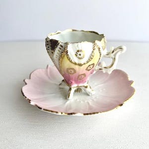 May include: A vintage teacup and saucer set. The teacup features a gold-trimmed rim and handle, with a floral design in shades of pink, yellow, and brown. The saucer is shaped like a flower, with a pink and white gradient and gold trim.