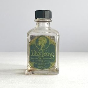 May include: A small, clear glass bottle with a black cap. The bottle has a green and gold label with the text "Day-tone Beauty Preparations". The label features a silhouette of a woman's profile. The bottle is likely vintage.