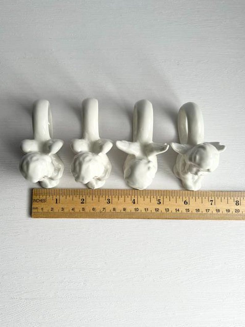 K&ouml;nnte beinhalten: Vier wei&szlig;e Serviettenringe aus Keramik, jeweils mit einem einzigartigen Tierdesign. Die Serviettenringe sind in einer Reihe angeordnet, darunter ein Holzlineal. Das Lineal misst bis zu 20 cm.