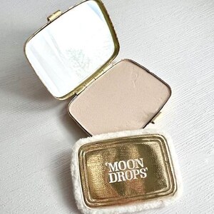 May include: A rectangular gold-tone compact case with a hinged lid and an interior mirror. The lid features the text 'MOON DROPS' in white lettering and is surrounded by a white faux fur trim. The compact contains a pressed beige powder.