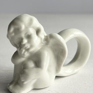 K&ouml;nnte beinhalten: Ein wei&szlig;er Serviettenring aus Porzellan in Form eines Cherubs. Der Cherub sitzt, den Kopf auf die Hand gest&uuml;tzt, und der Ring bildet eine Schlaufe dahinter. Das St&uuml;ck hat eine glatte, gl&auml;nzende Oberfl&auml;che und dient wahrscheinlich dekorativen Zwecken.