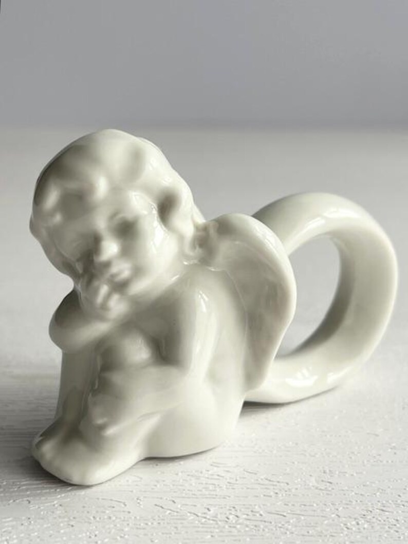 K&ouml;nnte beinhalten: Ein wei&szlig;er Serviettenring aus Porzellan in Form eines Cherubs. Der Cherub sitzt, den Kopf auf die Hand gest&uuml;tzt, und der Ring bildet eine Schlaufe dahinter. Das St&uuml;ck hat eine glatte, gl&auml;nzende Oberfl&auml;che und dient wahrscheinlich dekorativen Zwecken.
