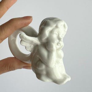 K&ouml;nnte beinhalten: Ein wei&szlig;er Serviettenring aus Keramik in Form eines Engels. Der Engel ist in sitzender Position dargestellt, den Kopf auf die Hand gest&uuml;tzt. Der Serviettenring hat eine Schlaufe zum Halten einer Serviette. Der Engel hat Fl&uuml;gel und tr&auml;gt ein drapiertes Gewand.
