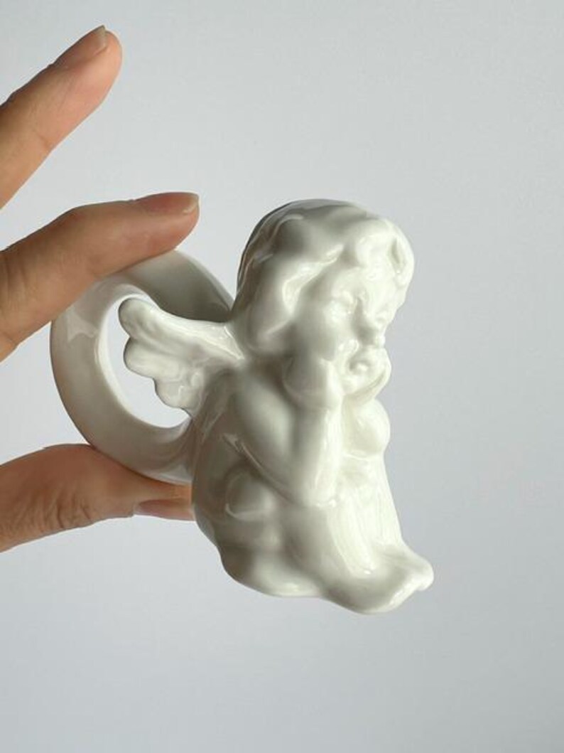 K&ouml;nnte beinhalten: Ein wei&szlig;er Serviettenring aus Keramik in Form eines Engels. Der Engel ist in sitzender Position dargestellt, den Kopf auf die Hand gest&uuml;tzt. Der Serviettenring hat eine Schlaufe zum Halten einer Serviette. Der Engel hat Fl&uuml;gel und tr&auml;gt ein drapiertes Gewand.